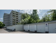 Mehrstöckiges Wohngebäude in grauer Plattenbauweise im Hintergrund, umgeben von Bäumen. Im Vordergrund befindet sich eine Reihe von weiß gestrichenen Garagen mit geschlossenen Toren auf einem gepflasterten Hof.