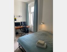 Schlafzimmer mit einem Doppelbett, bedeckt von einer grünlichen Bettdecke, und einem Stapel Handtücher darauf. Im Hintergrund ein Schreibtisch mit Computer und Stuhl vor einem hohen Fenster mit hellgrünen Vorhängen; an der Wand eine Wandlampe und ein Heizkörper.