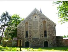 Steinbau einer historischen Kirche mit schrägem Satteldach, drei länglichen Spitzbogenfenstern und einer großen Inschrift auf der Vorderseite. Umgeben von Bäumen und einer Wiese, mit einem kleinen Mauervorsprung links im Bild.