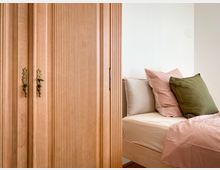 Holzschrank mit vertikalen Rillen und dekorativen Messinggriffen neben einem Bett mit pastellrosa, olivgrünen und beigen Kissen, in einem hellen, minimalistisch eingerichteten Raum.