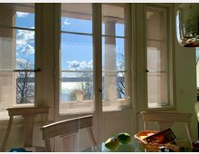 Heller Innenraum mit Blick durch gläserne Doppeltüren auf eine Terrasse mit Säulen, dahinter ein See mit Reflexionen des Sonnenlichts und kahlen Bäumen. Im Vordergrund ein Glastisch mit einem Obstkorb und dekorativen Stühlen.