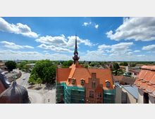 Blick auf ein historisches Gebäude mit einer Turmuhr und spitzem Turmdach, umgeben von roten Ziegeldächern und Gerüsten. Im Hintergrund sind eine Kleinstadtlandschaft, grüne Bäume sowie Windkraftanlagen unter einem blauen Himmel mit weißen Wolken zu sehen.