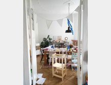 A cluttered dining area with a wooden table surrounded by mismatched chairs, a children's step stool in the foreground, and a rug covering wooden parquet flooring. The room is decorated with white triangular bunting, potted plants, framed wall art, and a black hanging light fixture.