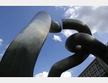 Großformatige, abstrakte Metallskulptur mit verschlungenen Röhren, die in den Himmel ragen, vor einem Hintergrund aus blauem Himmel mit Wolken und einem angrenzenden Gebäude.