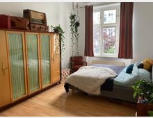 Ein Schlafzimmer mit Holzboden, einem großen Kleiderschrank mit Glasfronten, einem Bett mit mehreren Kissen und Decken, sowie einem Fenster mit braunen Vorhängen. Auf dem Schrank stehen ein altes Radio, Reisetaschen und Pflanzen; weitere Zimmerpflanzen und ein Sessel ergänzen die Einrichtung.