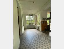 Ein heller Korridor mit großen Fenstern, die viel Tageslicht hereinlassen. Der Boden ist mit sechseckigen Fliesen ausgelegt, die Decke zeigt eine schlichte Struktur, und ein Kronleuchter hängt in der Mitte; an einer Wand stehen Pflanzen und Bilder zur Dekoration.