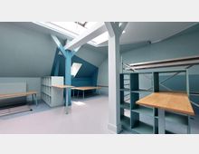 Ein heller Dachbodenraum mit blauen Wänden, mehreren Holztischen, offenen Regalen und einer sichtbaren Dachschräge. Das Tageslicht fällt durch Dachfenster, und eine Metallleiter sowie Baugerüste sind vorhanden.