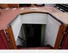 Treppenabgang in das Innere eines Schiffs mit einer geöffneten weißen Tür und Holzgeländer. Der Eingang ist von einer weißen Rahmenkonstruktion umgeben, und der Boden des Decks ist rot gestrichen.