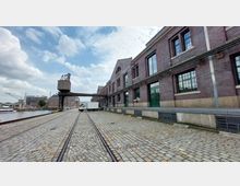 Backstein-Gebäude eines historischen Hafenareals mit großen Fenstern und grünen Türen, daneben ein Kopfsteinpflasterweg mit alten Gleisen. Im Hintergrund erkennbar ein Kran und weitere Hafengebäude an einem Wasserkanal.