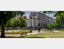 Wohngebiet mit einem mehrstöckigen Wohngebäude mit Balkonen und einem Eckgebäude mit rotem Dach und Arkaden im Erdgeschoss. Im Vordergrund ist eine Grünfläche mit Bäumen, einer Sitzbank und einem Radfahrer zu sehen.