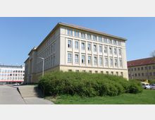 Gelbes, vierstöckiges Gebäude im klassischen Stil mit zahlreichen rechteckigen Fenstern und einem roten Satteldach, umgeben von grünem Strauchwerk und einem asphaltierten Weg mit Treppenstufen im Vordergrund. Im Hintergrund sind weitere moderne und klassische Gebäude sichtbar.