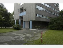 Wohnblock aus der Nachkriegszeit mit grauer Betonfassade, großen Fenstern und blauen Pfeilern im Erdgeschoss. Im Vordergrund sind ein gepflasterter Weg, grüne Rasenflächen und Büsche zu sehen.