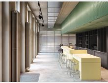 Moderner Innenraum einer Kantine oder Bar mit einem langen, hellgelben Tresen und silbernen Hockern auf Betonboden. Links stehen hohe Fenster mit vertikalen Heizkörpern, rechts befinden sich dunkle Regale und eine grün strukturierte Deckenverkleidung.