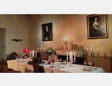 Historischer Speisesaal mit einer langen, festlich gedeckten Tafel, geschmückt mit Kerzenleuchtern, Geschirr und Dekoration. Im Hintergrund stehen ein Kamin mit Zierobjekten und Gemälde an den Wänden.