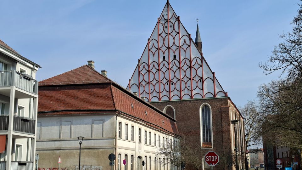 Straßenansicht eines Kirchengebäudes mit großer, dreieckiger Giebelfassade, die mit einem rot umrahmten Bogenmuster verziert ist, sowie einem seitlichen Backsteinbau mit hohen Rundbogenfenstern. Im Vordergrund stehen Wohnhäuser mit roten Ziegeldächern, Straßenlaternen und Verkehrsschildern, daneben Bäume entlang der Straße.