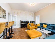 Modern eingerichtetes Wohnzimmer mit Fischgrät-Holzboden, samtigem Ecksofa in Petrol und gelben Sesseln samt Hocker um einen rechteckigen Couchtisch. An der Rückwand steht ein schwarzes Klavier vor einer hell gemusterten Akzentwand; links befindet sich ein schmaler Bartisch mit hohen Stühlen und zwei Türen führen in angrenzende Räume.