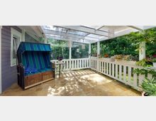 Überdachte Veranda mit weißen Holzgitterwänden, bepflanzten Blumenkästen und einem Strandkorb mit blau-grüner Polsterung; Steinboden und Glasdach sorgen für eine helle Atmosphäre, im Hintergrund grüne Vegetation.