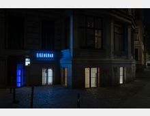 Außenansicht eines Gebäudeecks bei Nacht mit beleuchteten Fenstern und dem Leuchtschild „EIGENGRAU“ in blauer Schrift. Im Erdgeschoss sind vergitterte Fenster sowie eine blaue, beleuchtete Eingangstür zu sehen, die von Pflastersteinen und minimaler Straßenbeleuchtung umgeben sind.