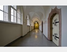 Langer, heller Korridor mit Spitzbogen-Gewölbedecke, großen Fenstern auf der linken Seite und verzierten Wandbildern sowie einer Tür mit aufwendiger schmiedeeiserner Verzierung auf der rechten Seite. Der Boden besteht aus grauem Material, und eine hängende Kugellampe ist im Hintergrund sichtbar.