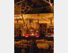 Ein rustikaler Innenbereich mit warmem Licht, geschmückt mit Lichterketten und dekorativen Elementen wie einem weißen, kahlen Baum. Im Vordergrund sind eine Bar mit roten Kerzen und Polsterhocker mit Fellüberzügen erkennbar, dahinter Fenster, Flaschen und weitere Einrichtungsdetails.