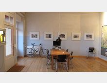 Ein heller Raum mit Holzboden, einem Holztisch und vier Stühlen. An der Wand hängen gerahmte Bilder, daneben steht ein Fahrrad, und eine Karte ist an der rechten Wand befestigt.