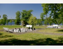 Grüne Parklandschaft mit großen Schachfeldern aus Stein und überdimensionierten Schachfiguren. Im Hintergrund sind Bäume, eine Uferpromenade und ein See zu sehen, umgeben von vereinzelten weißen Bänken.