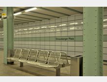 U-Bahn-Station mit gefliester Wand, auf der „Strausberger Platz“ steht, einer Sitzreihe aus Metallgittern und blassgrünen Stahlstützen. Die Beleuchtung besteht aus langen, rechteckigen Deckenleuchten.
