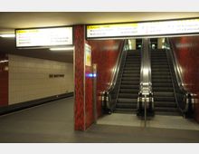 U-Bahnhof mit Innenansicht eines Bahnsteigs und einer Rolltreppe, die von roten Kachelwänden eingerahmt ist. Oben befinden sich gelb-weiße Hinweisschilder mit Ausgangs- und Streckeninformationen, und an der Wand ist der Schriftzug "Alt-Mariendorf" zu sehen.