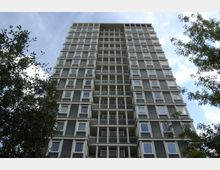 Ein modernes Hochhaus mit grauer Fassade, vielen gleichmäßig angeordneten Fenstern und Balkonen, umgeben von Bäumen im Vordergrund. Der Himmel im Hintergrund ist teilweise bewölkt.