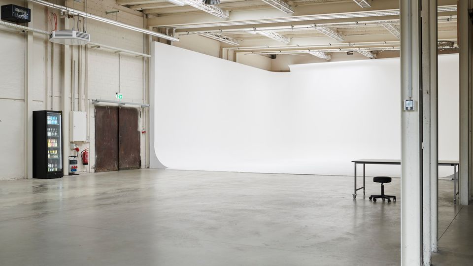 Large industrial studio space with a white cyclorama backdrop, concrete floor, and exposed ceiling beams with skylights and strip lighting. Along one wall are metal double doors, a vending machine, and visible pipes, with a table and stool in the foreground.