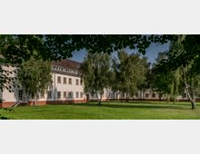 Zweistöckiges Gebäude mit heller Fassade und roten Klinkerfundamenten, großen rechteckigen Fenstern und einem Satteldach, umgeben von einer Wiese und zahlreichen Birkenbäumen im Vordergrund.