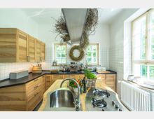 Helle Küche mit Holzmöbeln, einer Arbeitsplatte aus dunklem Material und einem zentralen Inselbereich mit Spüle und Gaskocher. Zwei Fenster mit weißen Rahmen lassen Tageslicht herein, während an der Wand weiße Fliesen und dekorative Elemente wie ein Wandkranz und Kräuterarrangements angebracht sind.