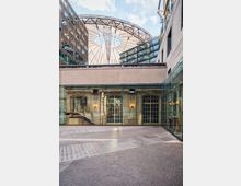 Innenhof mit gepflastertem Boden und verglaster Fassade, dahinter dekorative, historische Elemente wie verzierte Wände und große Fenster. Im Hintergrund ist ein modernes Dach mit radialer Konstruktion sowie umliegende moderne Gebäudefassaden zu sehen.