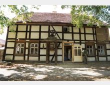 Fachwerkhaus mit weiß verputzten Wänden und dunklen Holzstreben, bestehend aus einem roten Ziegeldach und mehreren Fenstern. Im Vordergrund befinden sich Bäume, die Teile des Gebäudes verschatten, und eine offene Tür, die Einblick ins Innere bietet; eine Person steht rechts neben der Tür.