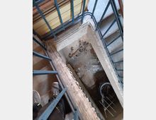 Blick in ein Treppenhaus mit blauen Metallgeländern. Der Boden des unteren Geschosses ist mit Bauschutt, Kabeln und Abfall bedeckt, während im Vordergrund ein Paar Beine und Schuhe auf einer Betontreppe zu sehen sind.