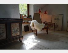 Gemütliche Ecke in einem Wohnzimmer mit hellem Holzfußboden, einem alten bemalten Holzschrank, einem Stuhl mit weißem Fell über der Lehne und rotem Lampenlicht. Im Hintergrund steht ein Fenster mit Blick ins Grüne, daneben Bücher, Dekorationsgegenstände und ein gerahmtes Bild an der Wand.