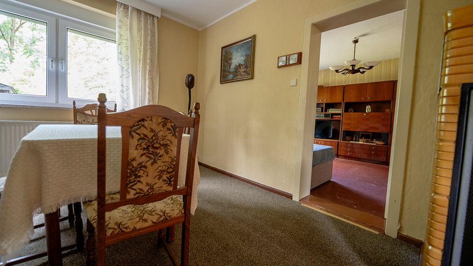 Blick in einen kleinen Essbereich mit Tisch und Polsterstühlen vor einem großen Fenster mit Gardinen. Durch eine offene Tür ist ein angrenzendes Wohnzimmer mit Holzschrankwand und Deckenleuchte zu sehen.