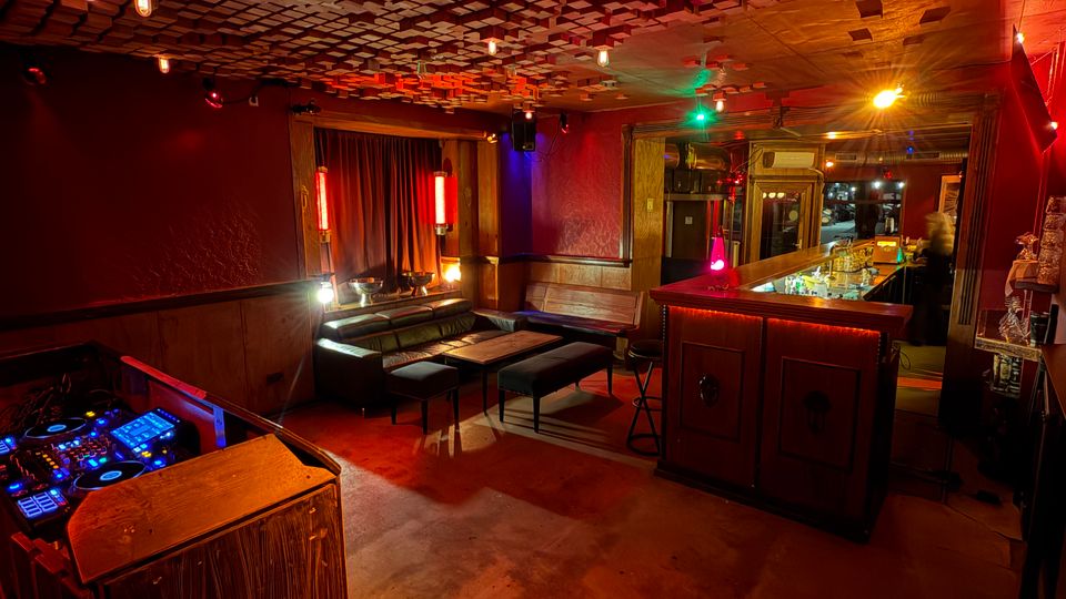 Innenraum einer Lounge-Bar mit dunkler Holzvertäfelung, Sitzbänken und niedrigen Tischen sowie einem Bartresen mit beleuchteten Flaschen. Auffällig sind die farbigen Lichtquellen und die strukturierte Decke mit vielen quadratischen Akustikelementen sowie ein DJ-Pult im Vordergrund.
