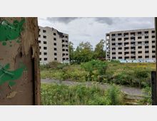 Verlassene Wohngebäude mit verblassten Fassaden stehen gegenüber einer verwilderten Grünfläche mit gelben Blumen und Gestrüpp; im Vordergrund ist eine beschädigte Wand mit grünen Graffiti sichtbar. Der Himmel ist bewölkt, und Bäume sind im Hintergrund zu erkennen.