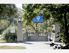 Eingang zur U-Bahn-Station „Hohenzollernplatz“ in Berlin, umgeben von Bäumen. Der steinerne Zugang mit Geländer und Laternen ist von einem blauen U-Bahn-Schild gekennzeichnet, im Hintergrund sind Gebäude und grüne Vegetation zu sehen.