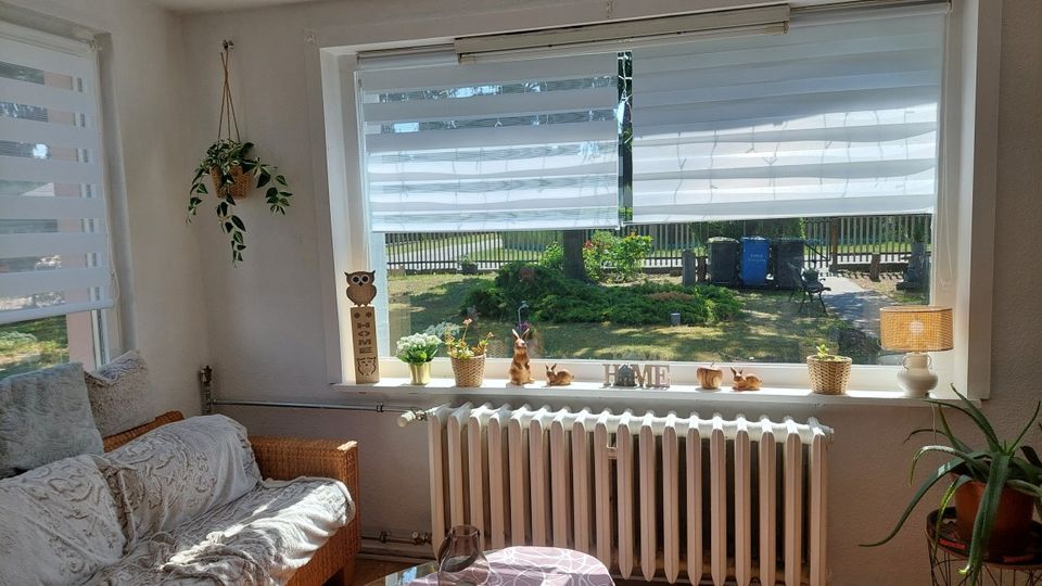 Helles Wohnzimmer mit großer Fensterfront und halb geöffneten Doppelrollos, breiter Fensterbank mit Pflanzen und Deko sowie einem Heizkörper darunter. Links steht ein Sofa neben einem Seitenfenster, rechts eine Stehlampe und Topfpflanze; durch das Fenster ist ein Gartenbereich sichtbar.