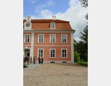 Barockes Gebäude mit rosa Fassade, weißen Fenstereinfassungen und einem roten Ziegeldach. Links ist ein Eingangsportal mit verziertem Türrahmen und Uhr; davor stehen mehrere Personen auf einem gepflasterten Hof.