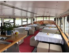 Innenraum eines Ausflugsbootes mit gedeckten Tischen, weißen Tischdecken und dekorativen Blumenvasen. Der Raum hat große Fenster mit Blick aufs Wasser, beige Sitzmöbel und rote Akzente wie einen Teppich; an der Decke hängen Dekorationen.