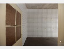 Ein leerer Raum mit weiß gefliesten Wänden und dunkelbraunem Boden. Links im Vordergrund ist eine beige Tür mit kleinen Fensterscheiben zu sehen.