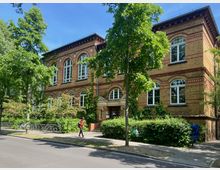 Zweistöckiges Backsteingebäude mit großen, weißen Bogenfenstern und Zierelementen aus roten Klinkersteinen, umgeben von Bäumen und Büschen. Vor dem Gebäude führt ein Gehweg mit Fahrrädern und einer Person, die vorbeigeht, entlang.