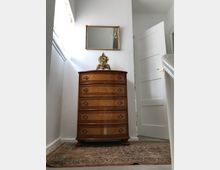 Holzkommode mit fünf Schubladen und dekorativen Griffen auf einem Teppich in einem hellen Raum; darüber hängt ein rechteckiger Wandspiegel mit goldgerahmtem Rahmen. Im Hintergrund ist eine geöffnete weiße Tür zu sehen.