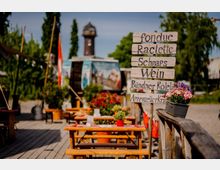 Holzterrasse eines Außenrestaurants mit Picknicktischen und rot-weiß karierten Tischdecken, dekoriert mit Blumen und Schweizer Flaggen. Im Vordergrund steht ein handgemaltes Holzschild mit Speise- und Getränkeangeboten; im Hintergrund sind Bäume und ein Turm zu sehen.