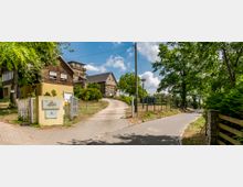 Zufahrt zu einem ländlichen Gebäudeensemble mit gemauerten und holzverkleideten Häusern, umgeben von Bäumen und Grünflächen. Ein gepflasterter Weg führt an einem Tor und einer Infotafel vorbei auf das Gelände, im Hintergrund eine historische Backsteinbauweise.