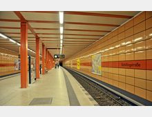 U-Bahn-Station Schillingstraße mit orange-braun gefliesten Wänden, orangefarbenen Stützen und einem gewölbten Bahnsteig neben den Gleisen. An den Wänden befinden sich Hinweisschilder und Werbung, die Station ist gut beleuchtet.