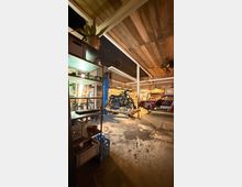 Innenansicht einer Werkstattgarage mit Holzbalkendecke, Betonboden und Werkregalen an der Seite. In der Mitte steht ein Motorrad auf einer Hebebühne, im Hintergrund ein teilweise zerlegtes Auto, beleuchtet von mehreren Hängelampen.